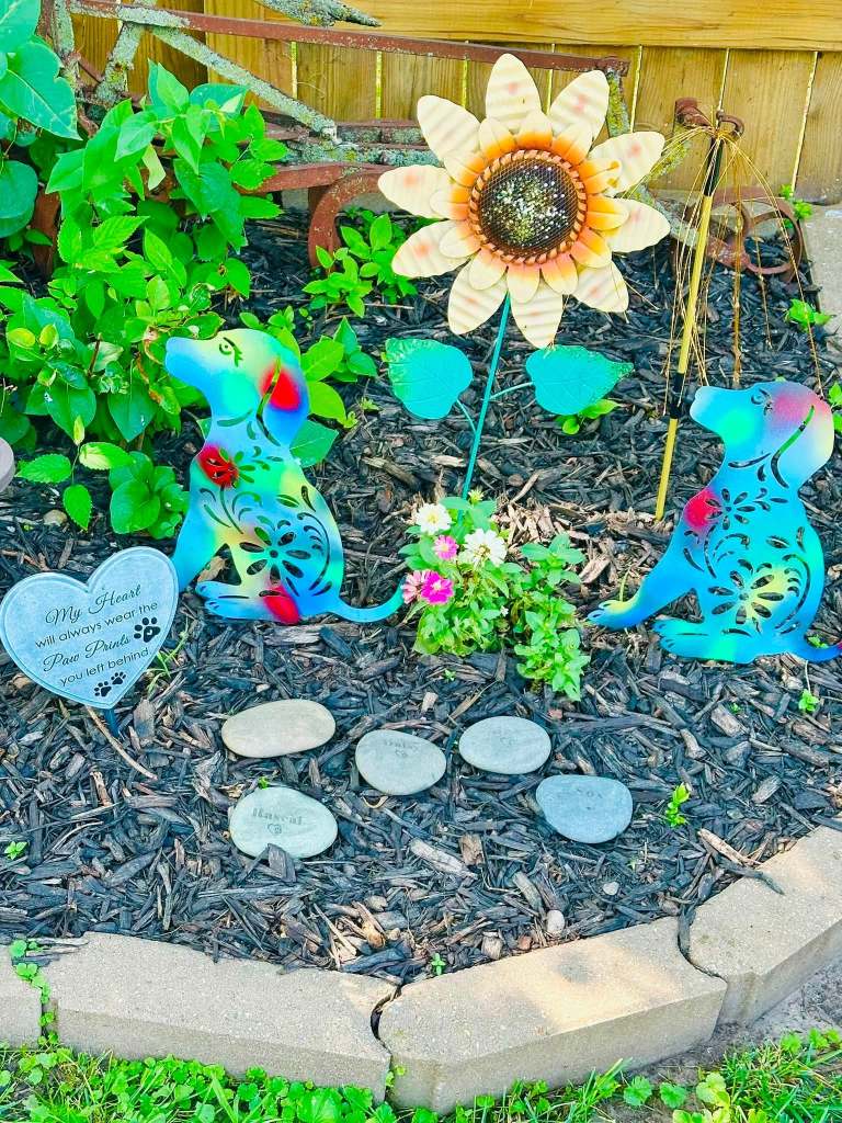 A decorative garden scene featuring two colorful metal dog sculptures, a large sunflower decoration, and a heart-shaped stone with an inscription. Small flowers and pebbles are arranged in a mulch bed, creating a heartfelt tribute.