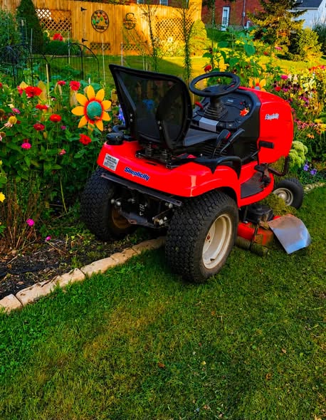 A red lawn mower parked along a flower garden, featuring colorful blooms and a decorative sunflower.