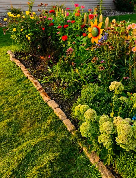 A colorful flower garden featuring various blooms, including yellow, red, and purple flowers, bordered by a stone pathway and green grass.