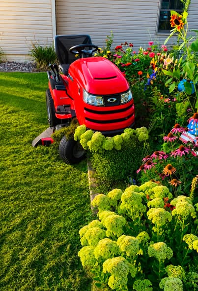 A bright red lawn tractor positioned next to a well-maintained flower garden featuring vibrant blossoms and neatly trimmed greenery.