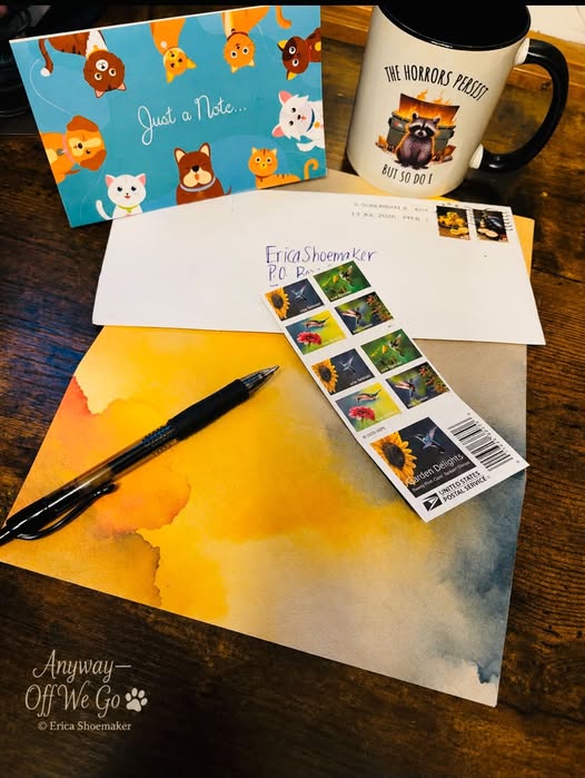 A colorful desk setup featuring a cat-themed notecard, a coffee mug with a humorous quote, an envelope addressed to 'Erica Shoemaker', a black pen, and a sheet of watercolor paper with sunflower postage stamps.