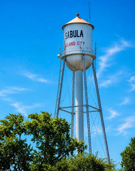 Water tower labeled 'SABULA ISLAND CITY' against a blue sky with some trees in the foreground.