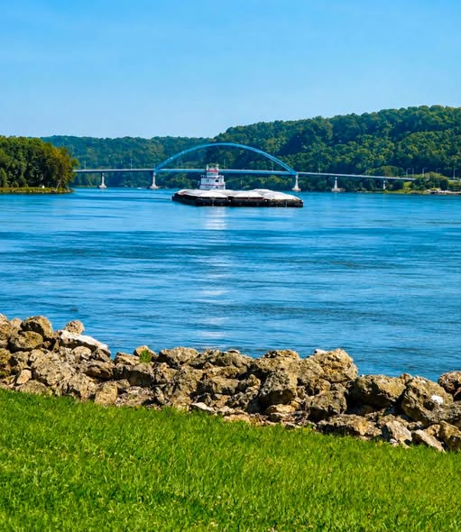 A river landscape featuring a barge traveling on calm water, with a bridge visible in the background and green hills along the shoreline.