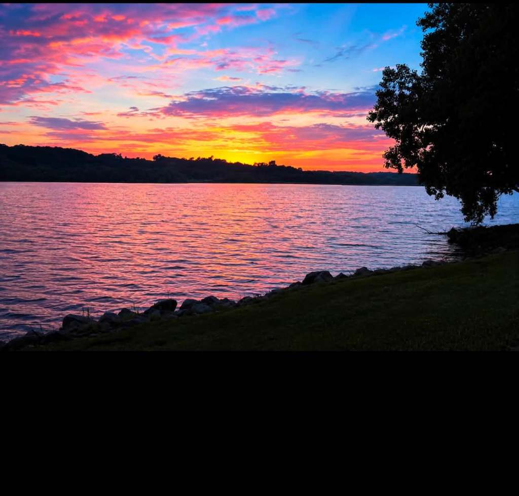 A serene sunset over a calm lake, featuring vibrant colors in the sky with hues of orange, pink, and blue, alongside a tree and rocky shoreline.
