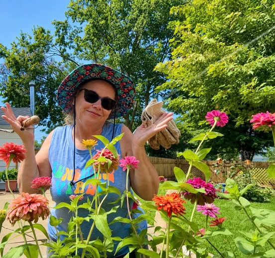 A woman in a garden surrounded by colorful flowers, wearing a wide-brimmed hat and sunglasses, holding gardening tools in one hand and gloves in the other, with a relaxed expression.