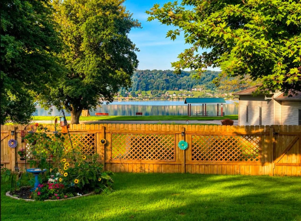 A scenic view from a backyard featuring a wooden fence, colorful flowers, and a grassy lawn, with a tranquil lake and hills in the background under a clear blue sky.