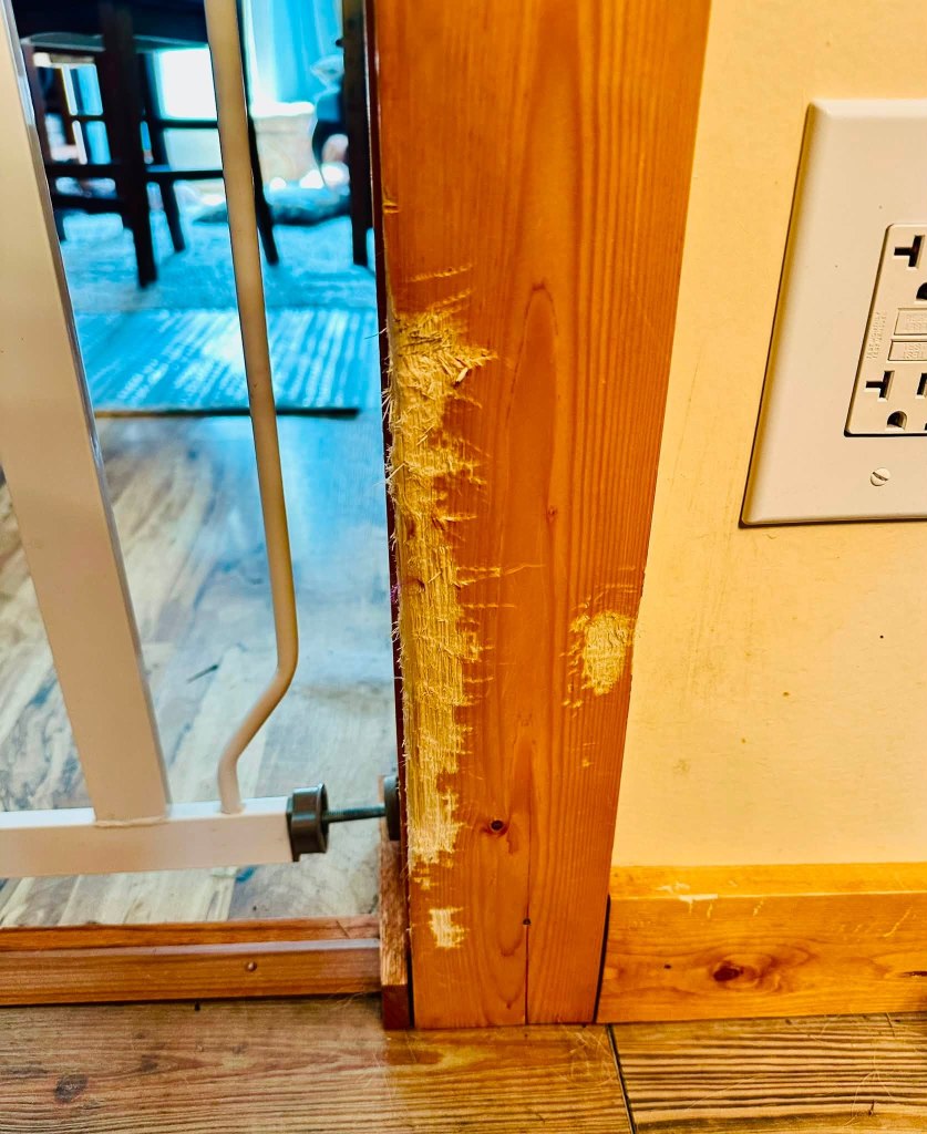 Close-up of a wooden door frame showing scratches and damage on the edge, with a wall outlet visible nearby.