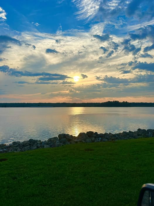 A serene sunset over a calm lake, with colorful clouds reflecting on the water's surface and a grassy shoreline in the foreground.