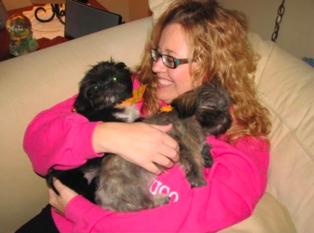 Woman sitting on a couch, smiling while holding two small dogs in her arms.
