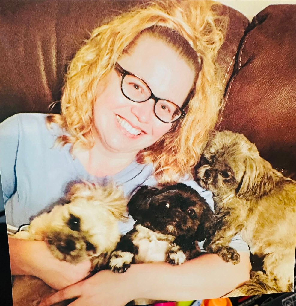 A smiling woman with curly blonde hair wearing glasses sits on a couch holding three small dogs, two with light fur and one black. The scene conveys a cozy and cheerful atmosphere.