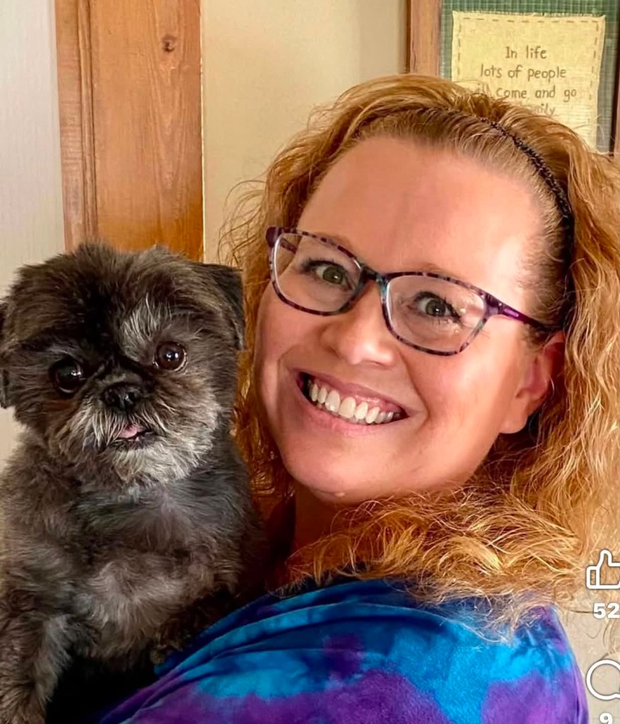 A woman with curly hair, wearing glasses, smiles while holding a small black dog with a slightly protruding tongue.